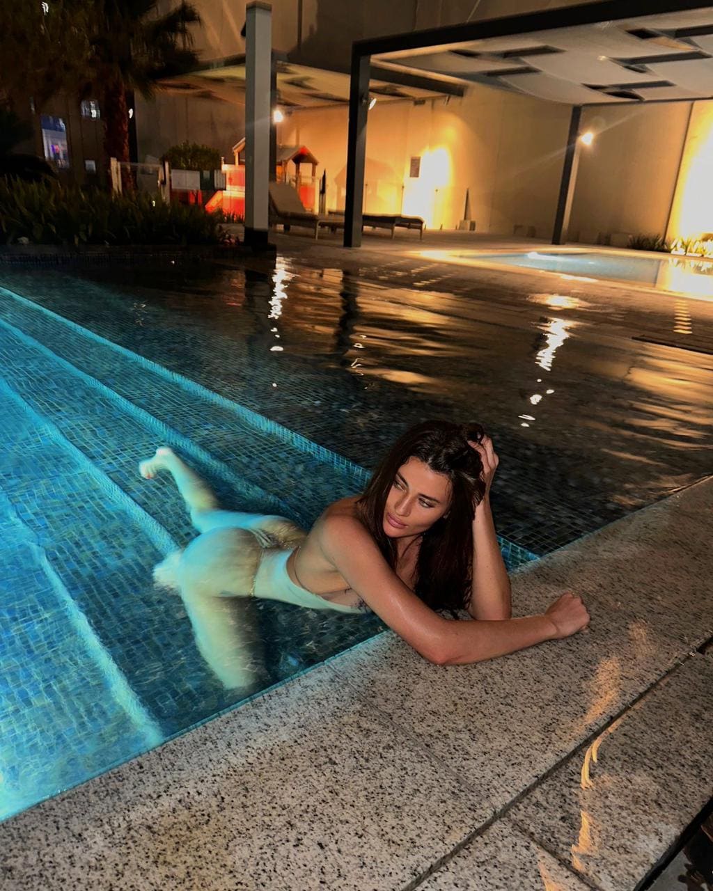 A woman rests her upper body on the stone edge of a tiled swimming pool at night, wearing a light-colored swimsuit.
