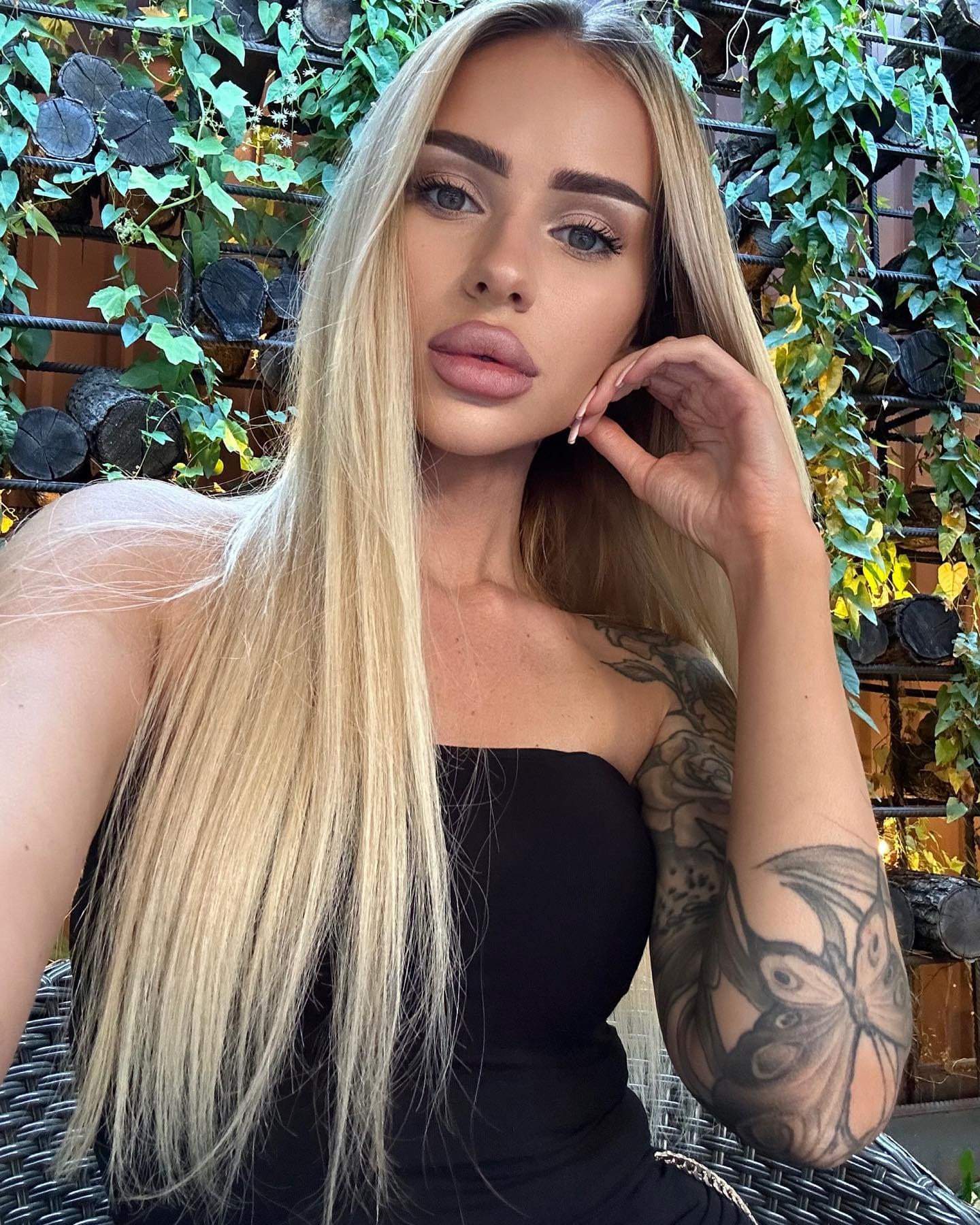 Alina wears a black strapless top, posing with a hand near her face to show a floral tattoo against a wall of green vines and logs.