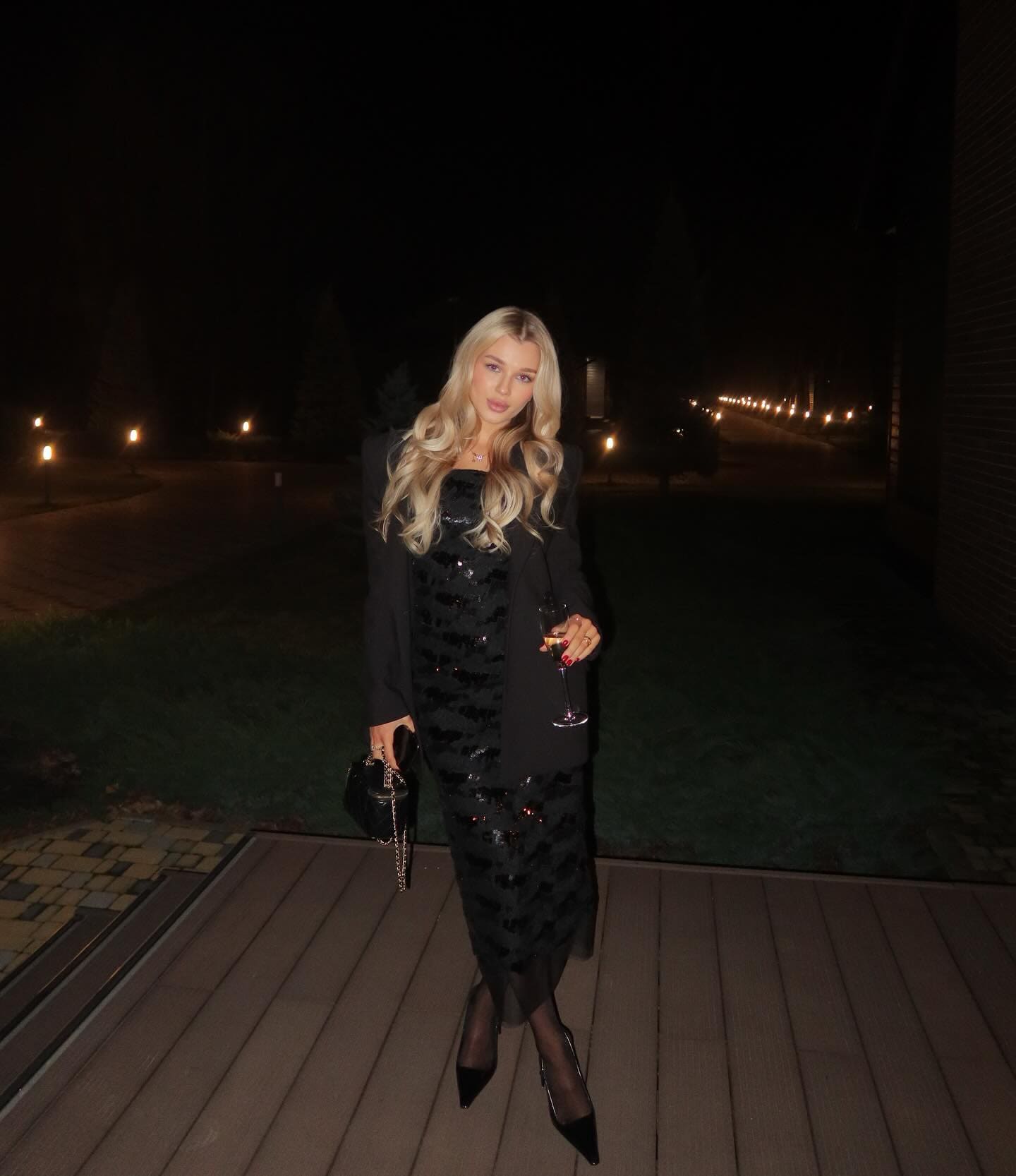 A woman in a black sequined dress and blazer stands on a deck at night, holding a wine glass and a small black handbag.