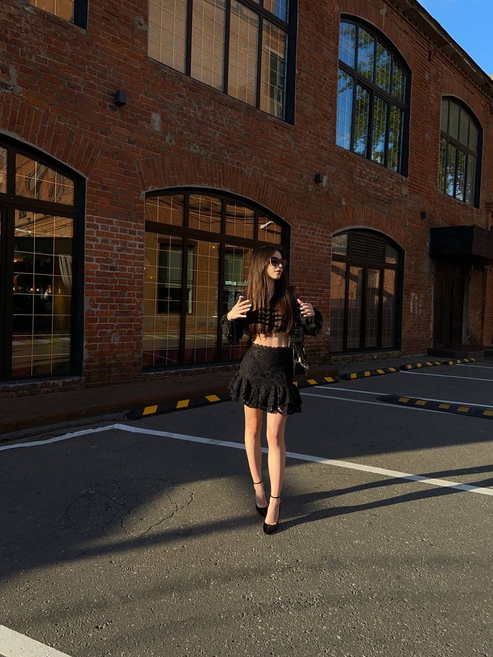 A woman in sunglasses, black lace crop top, and ruffled skirt stands in a parking lot near a brick building.