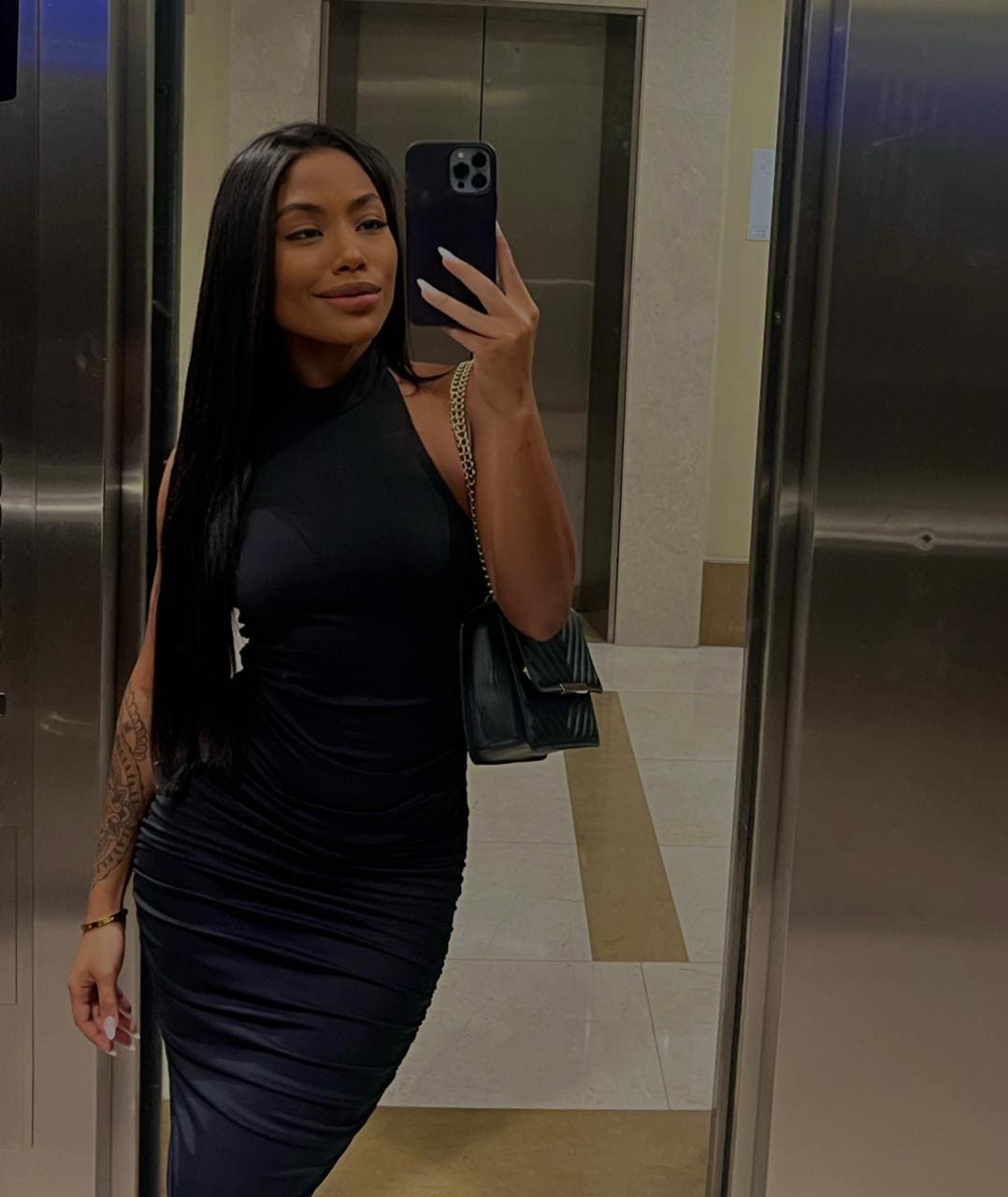 Elevator mirror selfie of a woman in a black halter dress with a tattoo on her forearm and quilted bag.