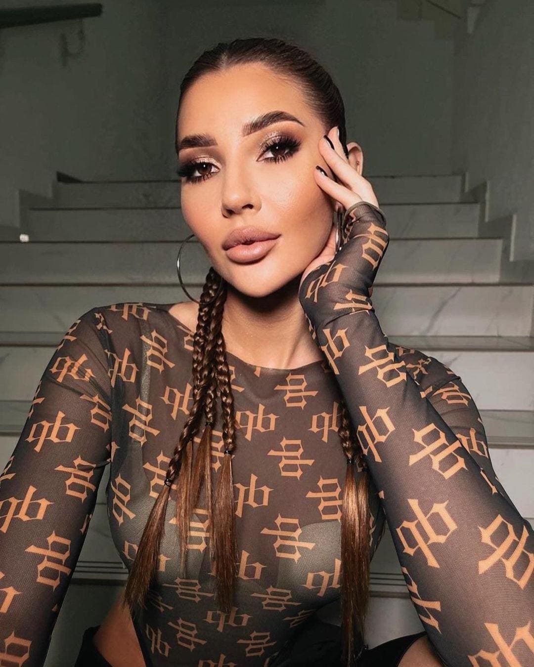 A woman with dark braids sits on concrete stairs, wearing a sheer gold-lettered top and hoop earrings, hand to cheek.