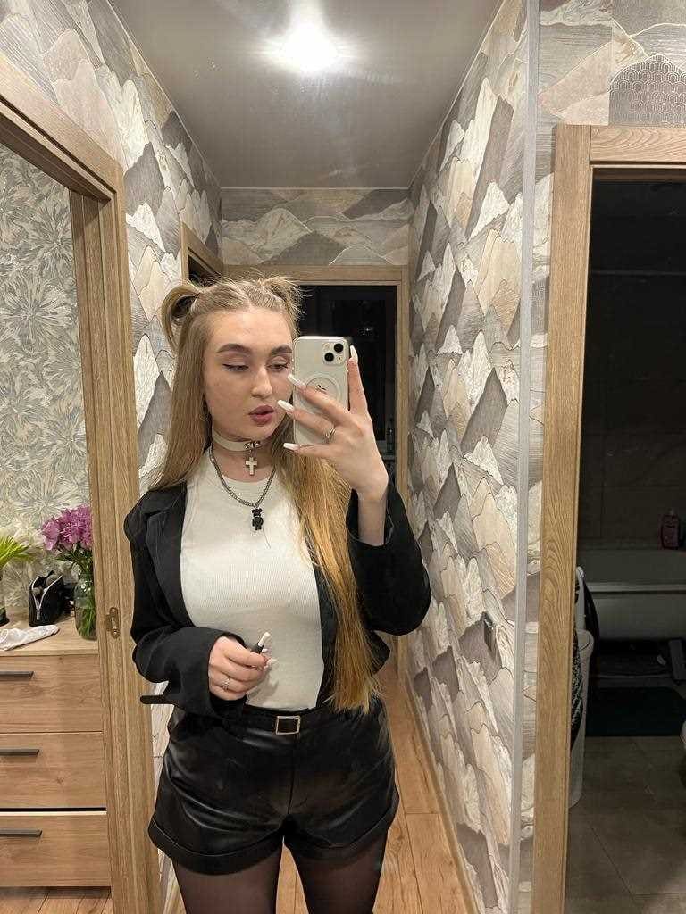 Alya takes a mirror selfie in a hallway, wearing a white top, black jacket, leather shorts, and tights with blonde pigtails.