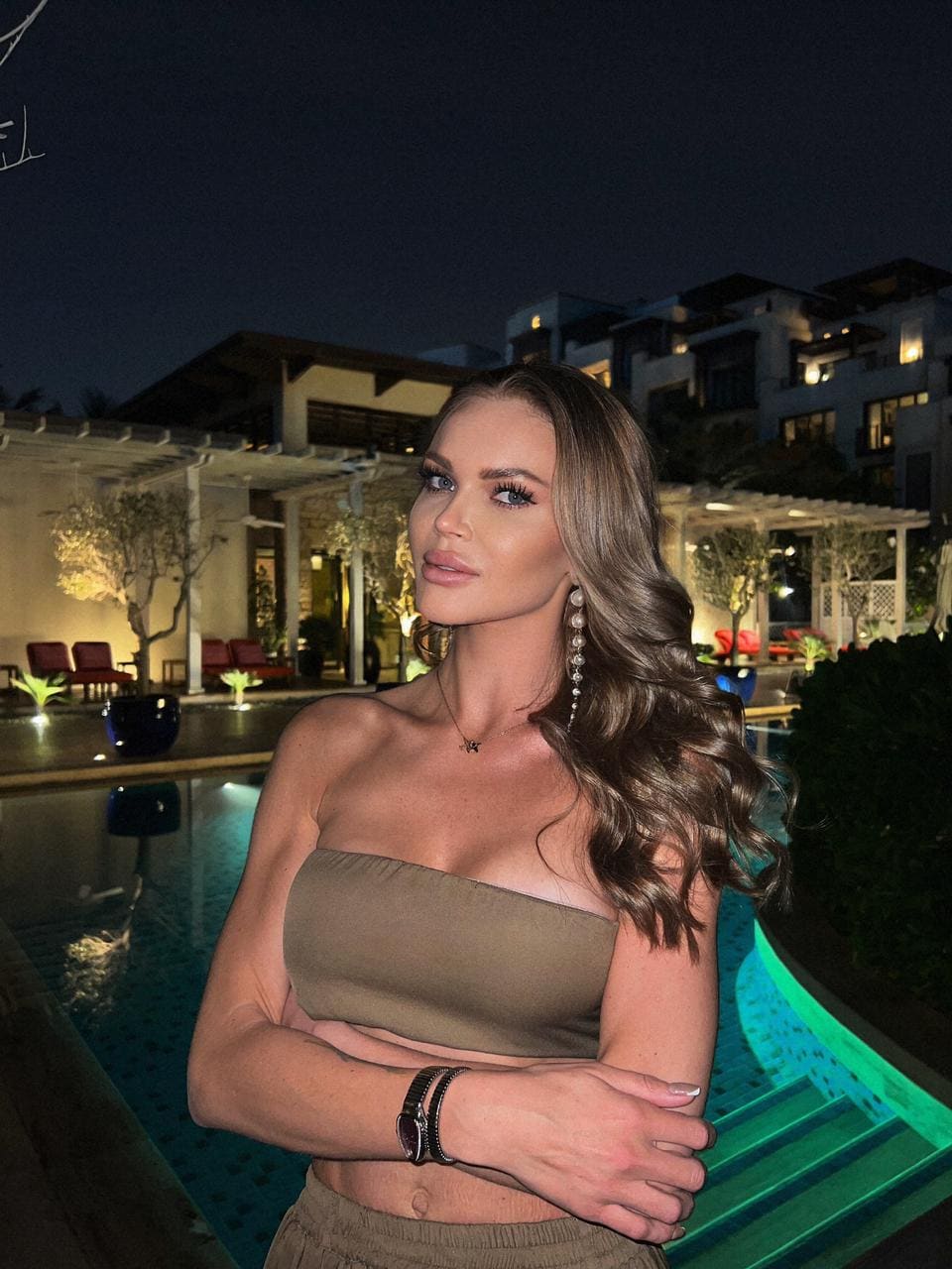 Woman with arms crossed by a lit pool at night, wearing an olive-green tube top and matching bottoms.