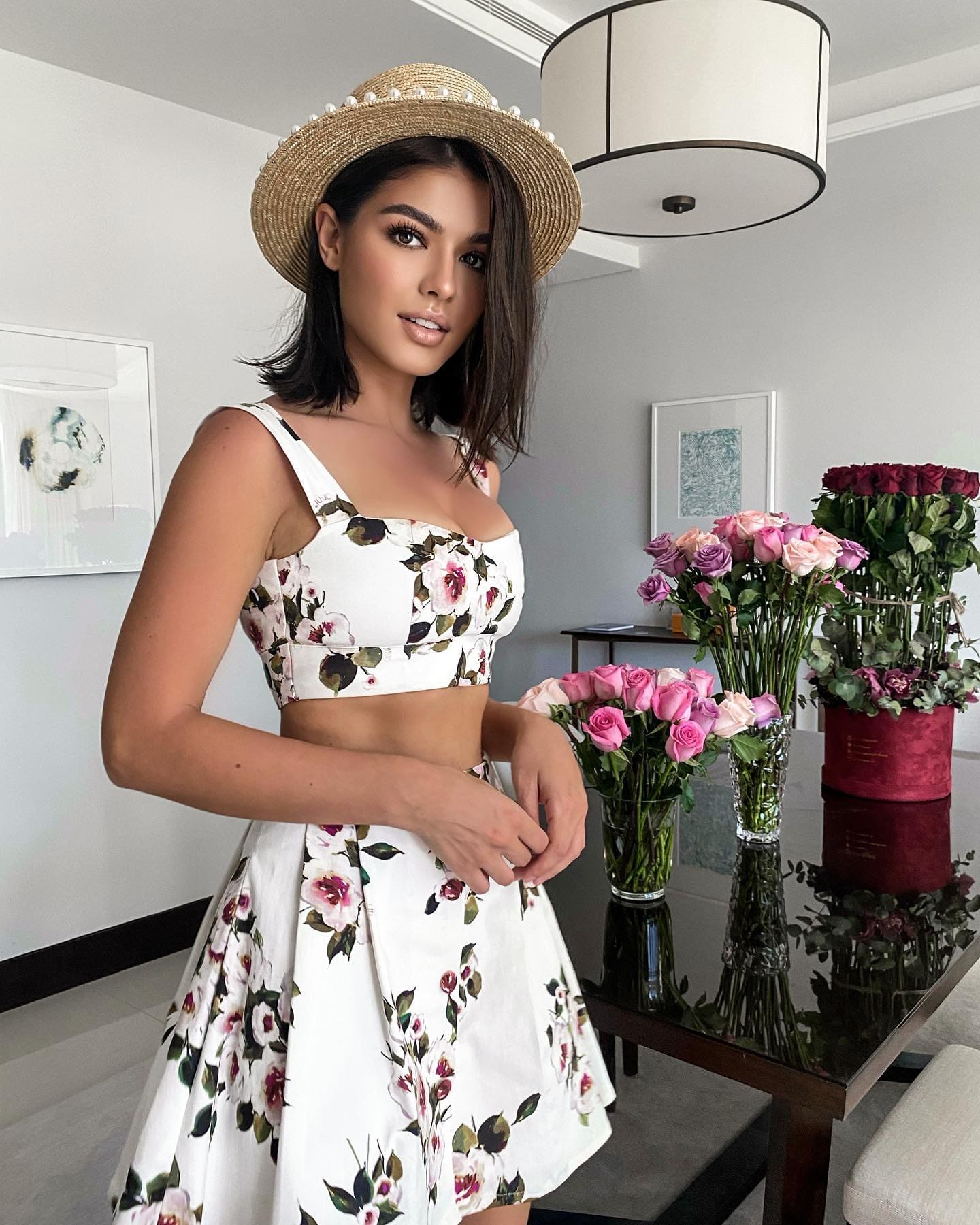 Eve stands indoors in a white floral crop top and skirt with a pearl-trimmed straw hat, roses in vases behind her.