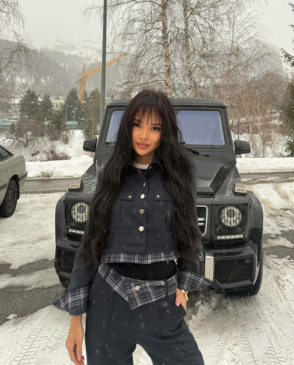 A woman with bangs wears a dark jacket with plaid cuffs, standing before a black SUV in a snowy landscape with bare trees.