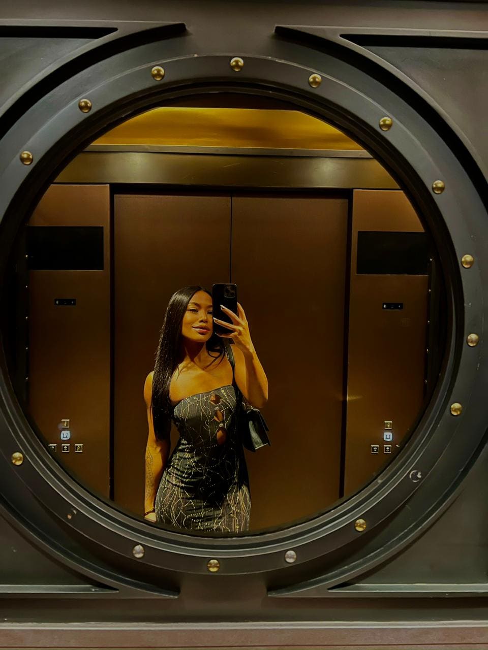 Mirror selfie through an elevator porthole window of a woman in a strapless patterned dress holding a phone.