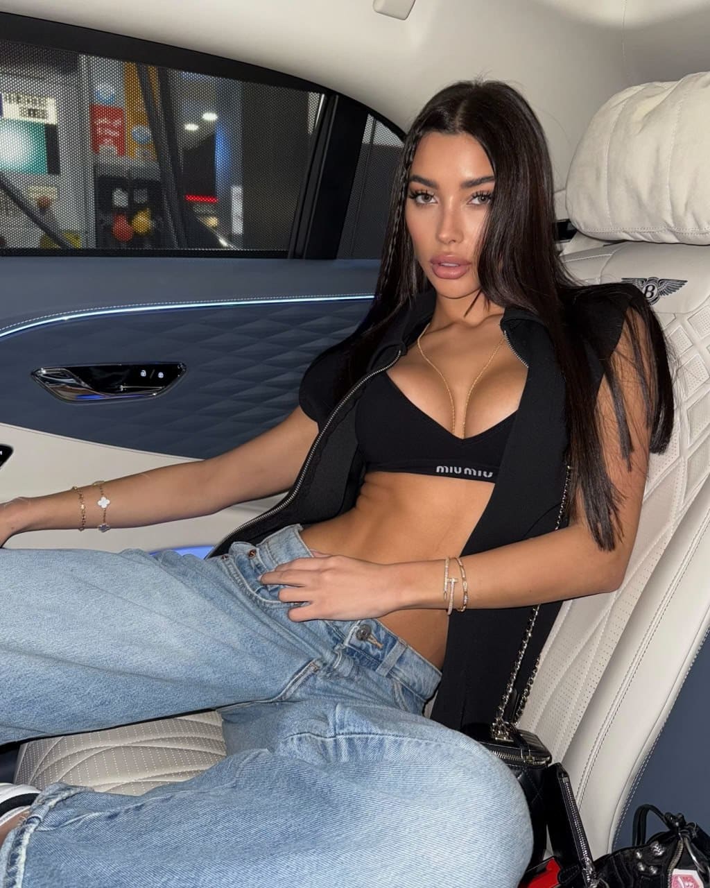 Seated in a car's back seat, a woman wears a black cropped zip-up jacket over a sports bra and light-wash jeans, hand on her lap.