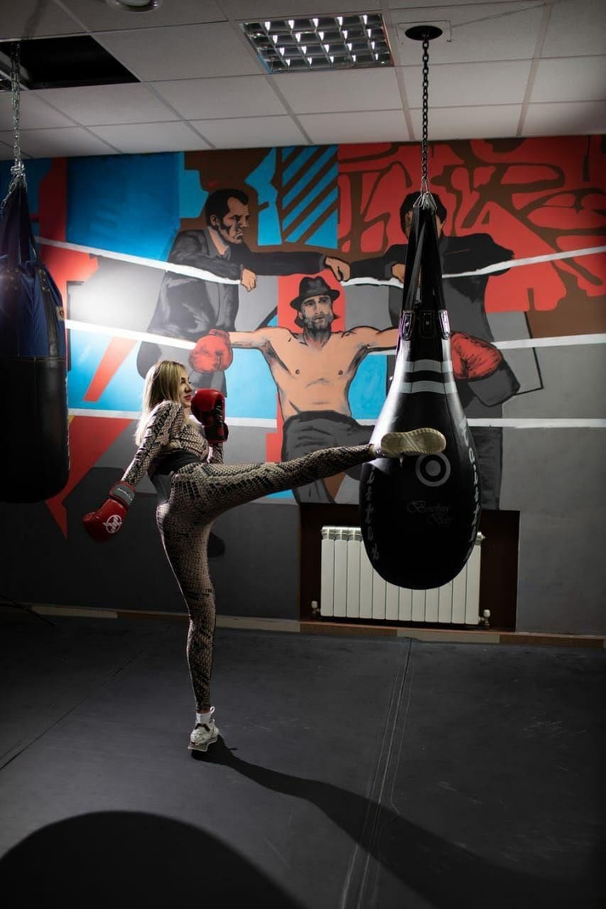A blonde woman in patterned athletic wear and red boxing gloves kicks a black punching bag in a gym featuring a colorful mural on the wall.