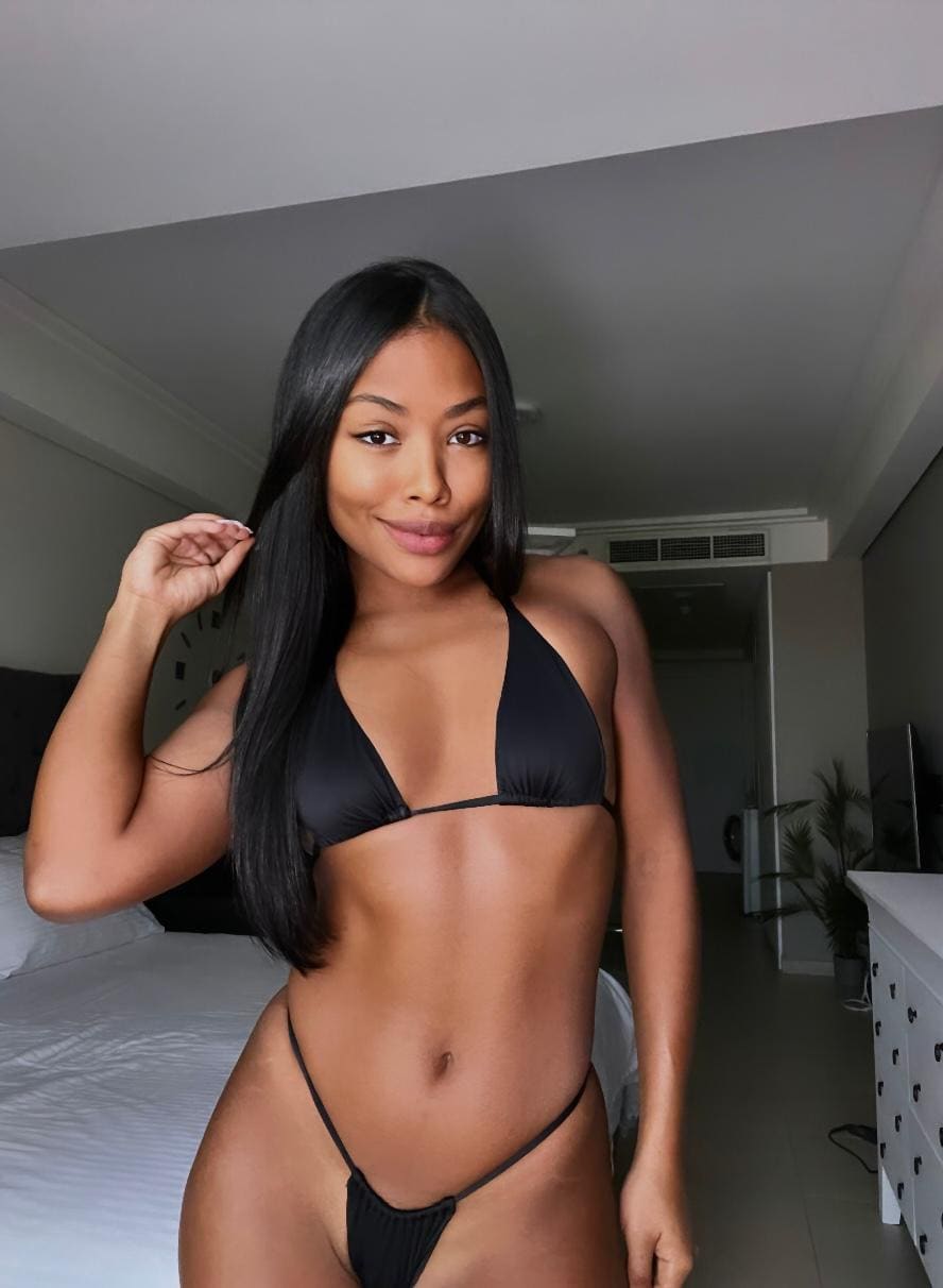 Woman in a black two-piece bikini stands in a bedroom with a bed and white dresser in the background.