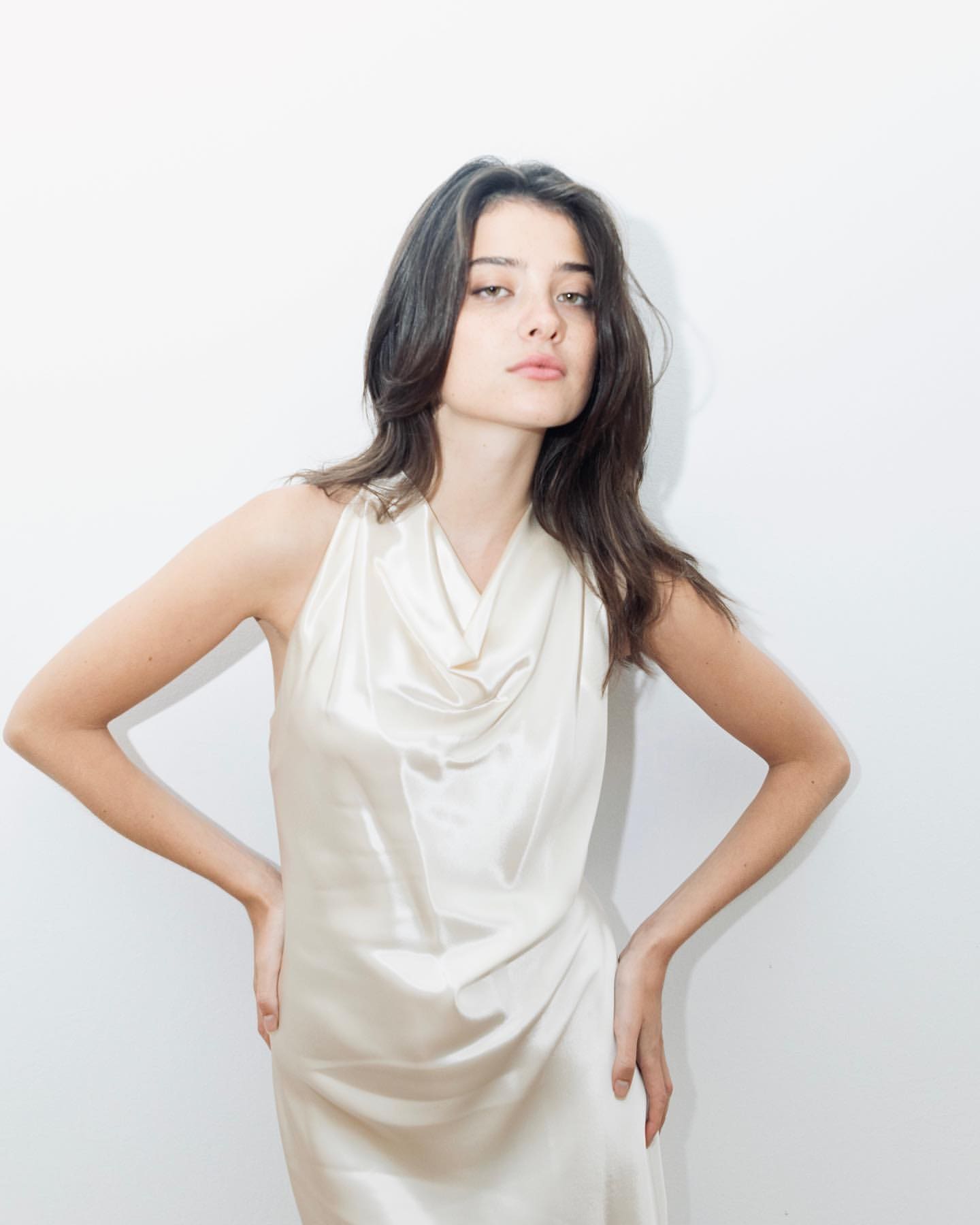 Blanca stands with hands on hips in a cream satin cowl-neck dress against a plain white background.