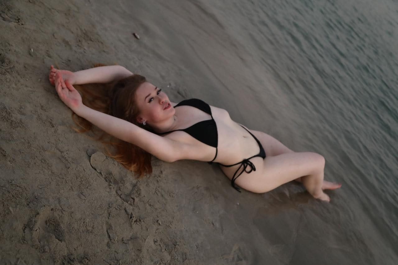 A red-haired woman lies on her side on wet sand in a black string bikini, arms raised above her head and knees bent.