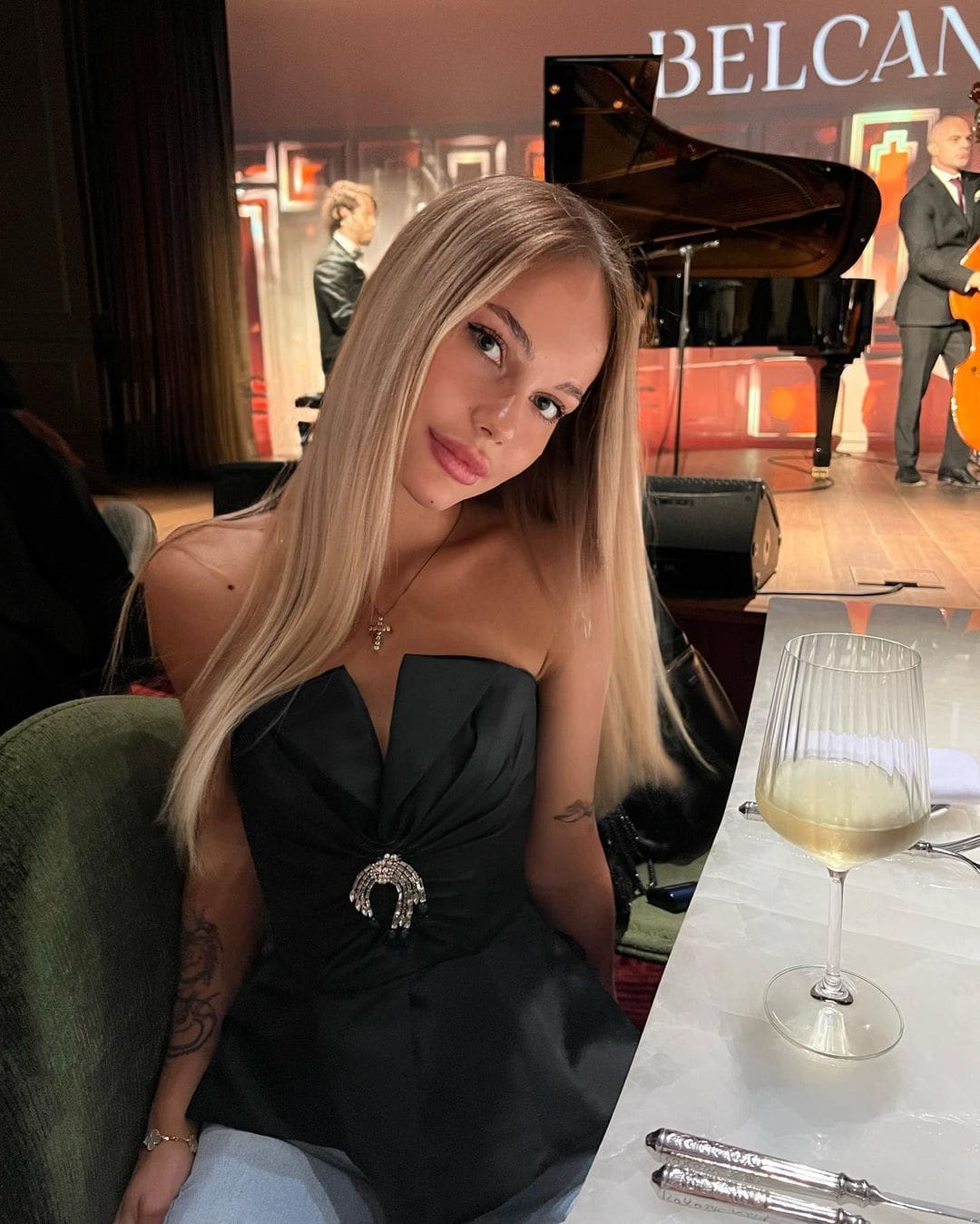 A blonde woman in a black strapless dress with a brooch sits at a table before a stage with a piano.