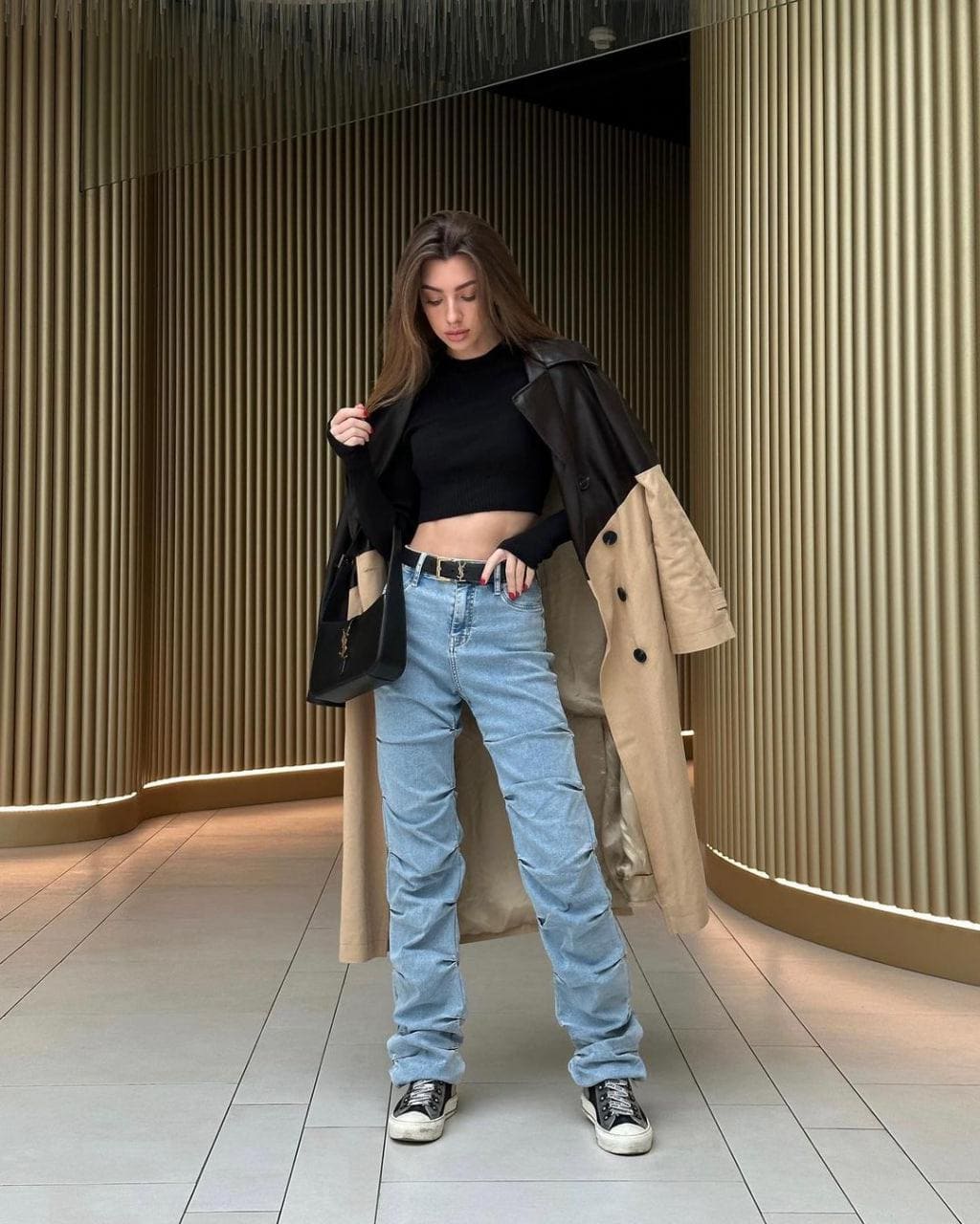 A woman stands in a tiled space with wooden slats, wearing a black cropped turtleneck, blue jeans, sneakers, and a tan coat with leather shoulders.
