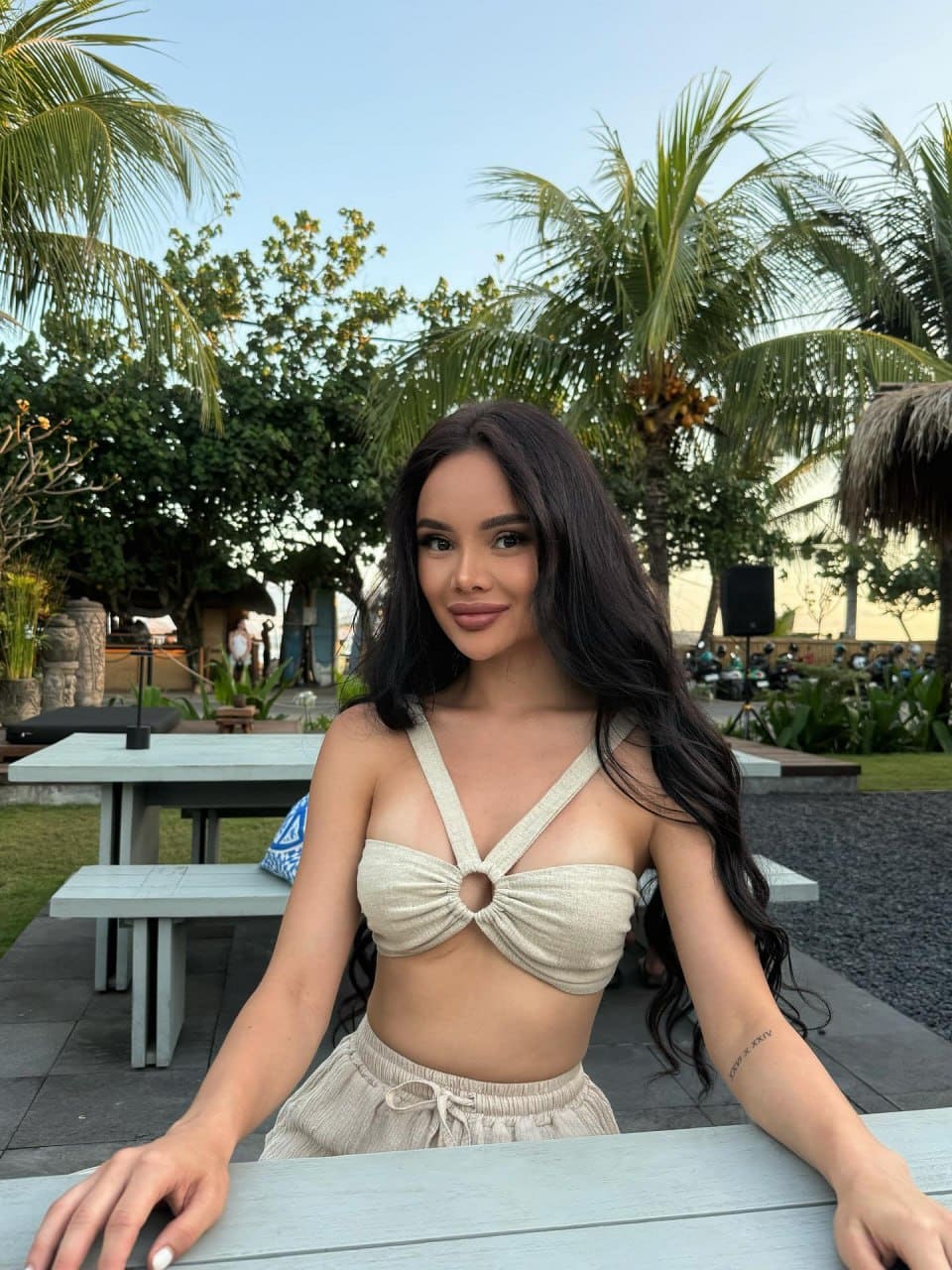 Zara sits at an outdoor table in a beige strappy crop top and drawstring pants, with palm trees and a garden patio behind her.