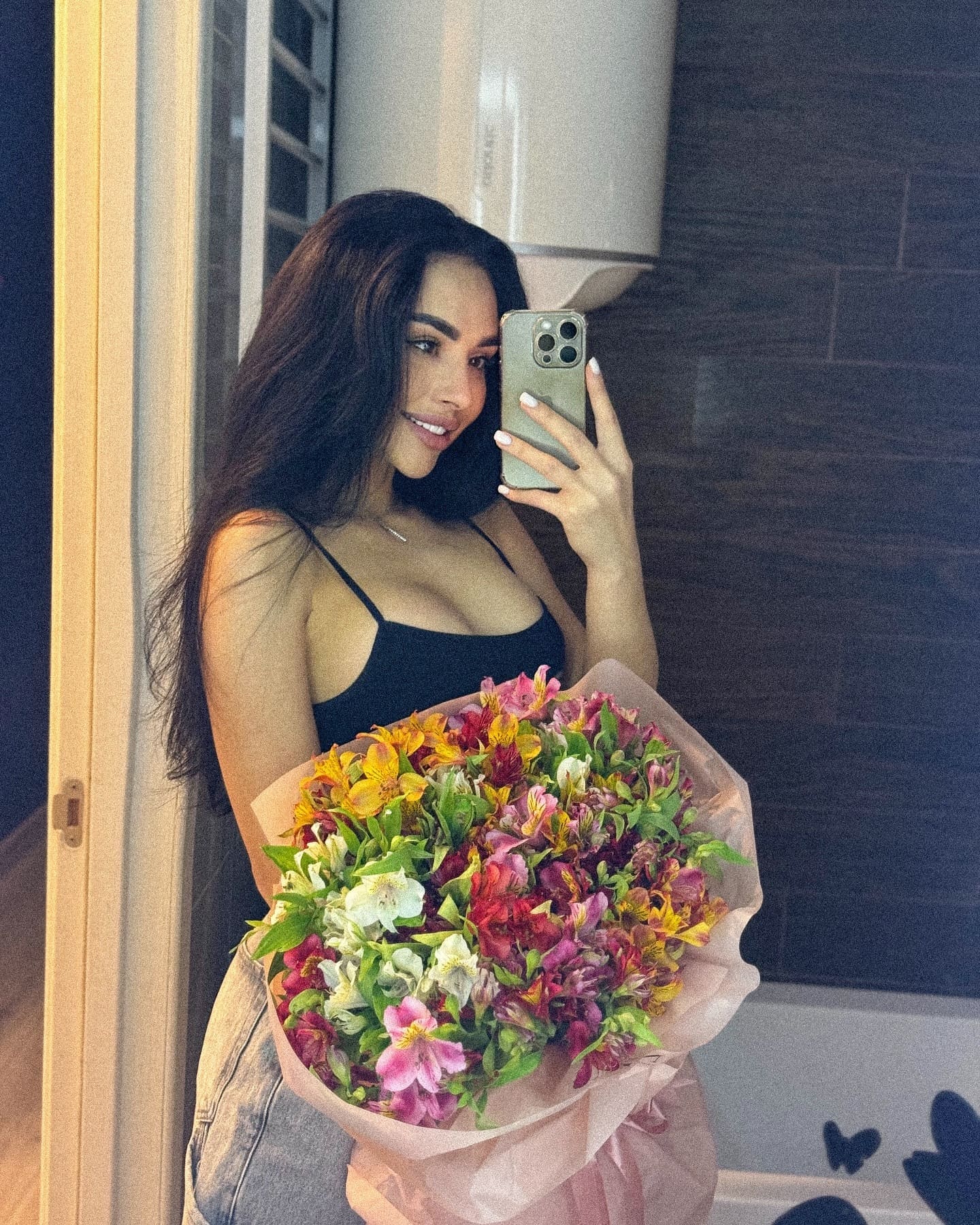 Mirror selfie showing a woman in a black top and jeans holding a large bouquet of mixed flowers in front of her.