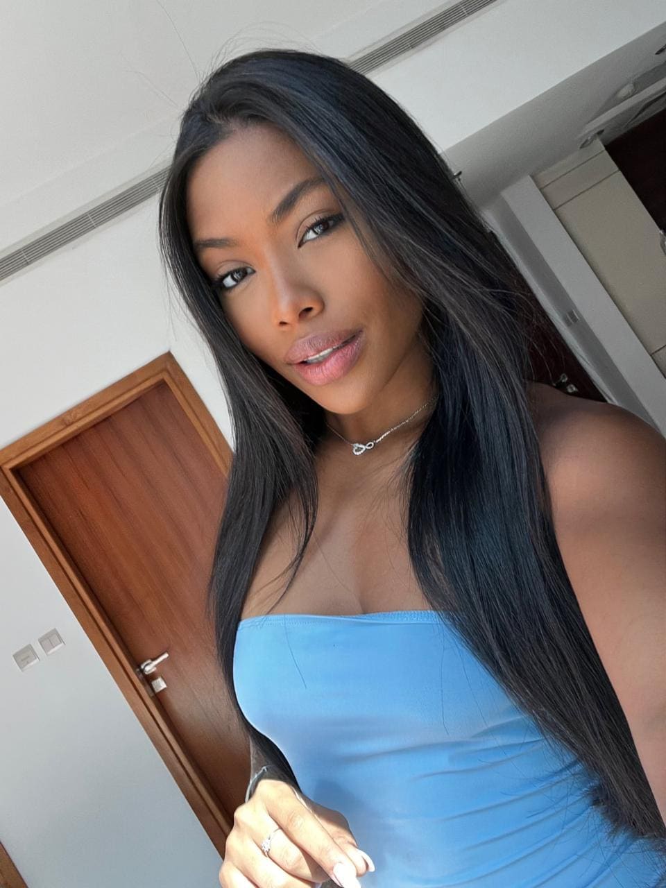 Naomi with long dark hair wears a light blue strapless top and necklace, posing indoors by a wooden door.