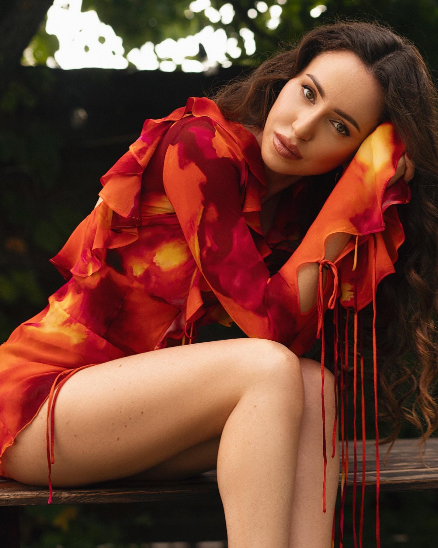 Lena sits on a wooden bench outdoors, wearing a short red and orange ruffled dress with cut-out sleeves, head resting on her hand and legs crossed.