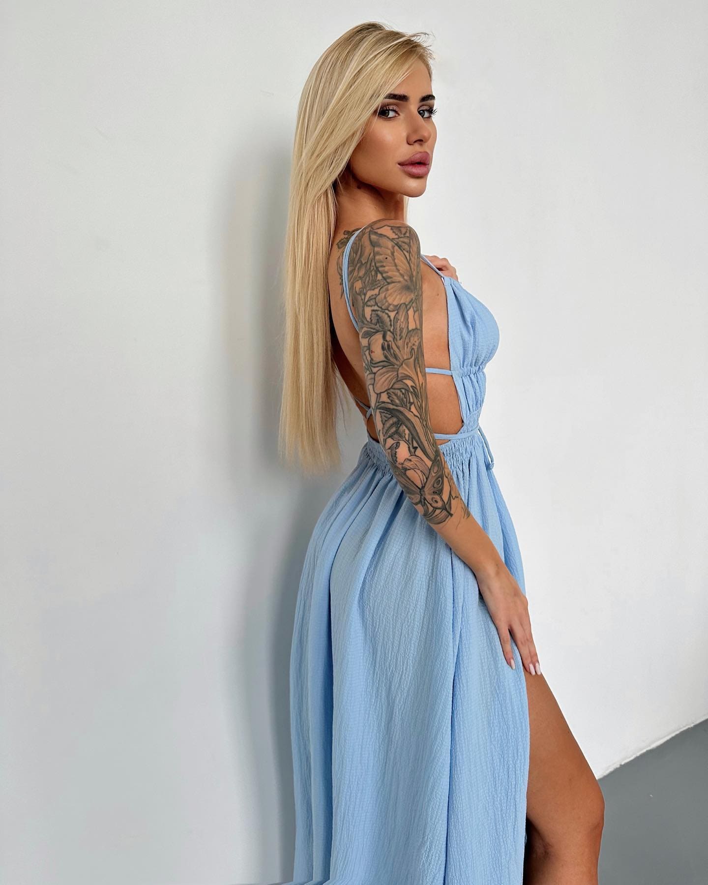 In profile against a white wall, she wears a light blue sleeveless dress with an open back and high slit, revealing a floral arm tattoo.
