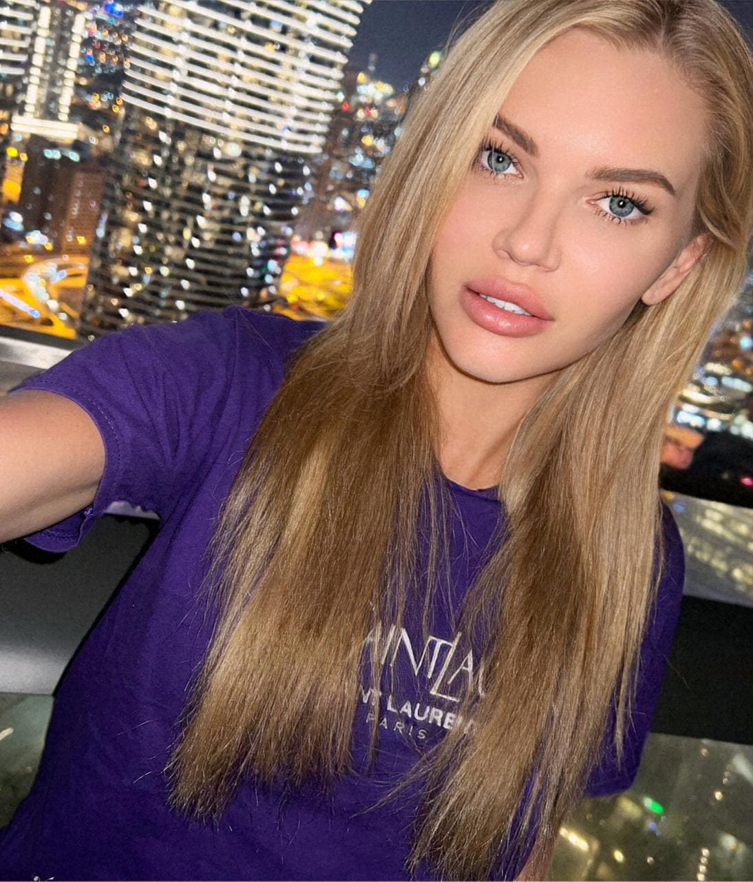 Blonde woman in a purple t-shirt taking a selfie with a night city skyline behind her.