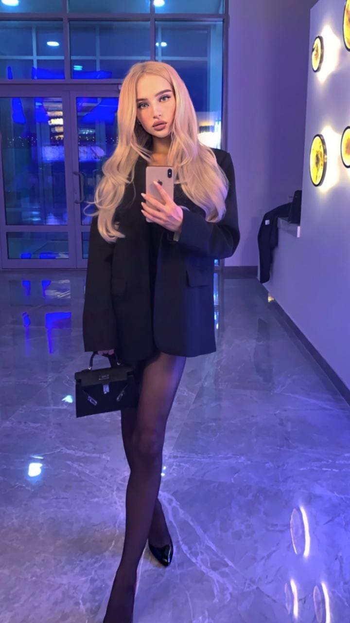 A woman in a black blazer and sheer tights takes a mirror selfie in a marble hallway, holding a bag.