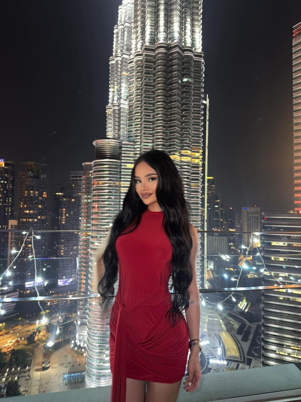 A woman in a red mini dress stands on a rooftop terrace at night, with a brightly lit skyscraper and city skyline in the background.