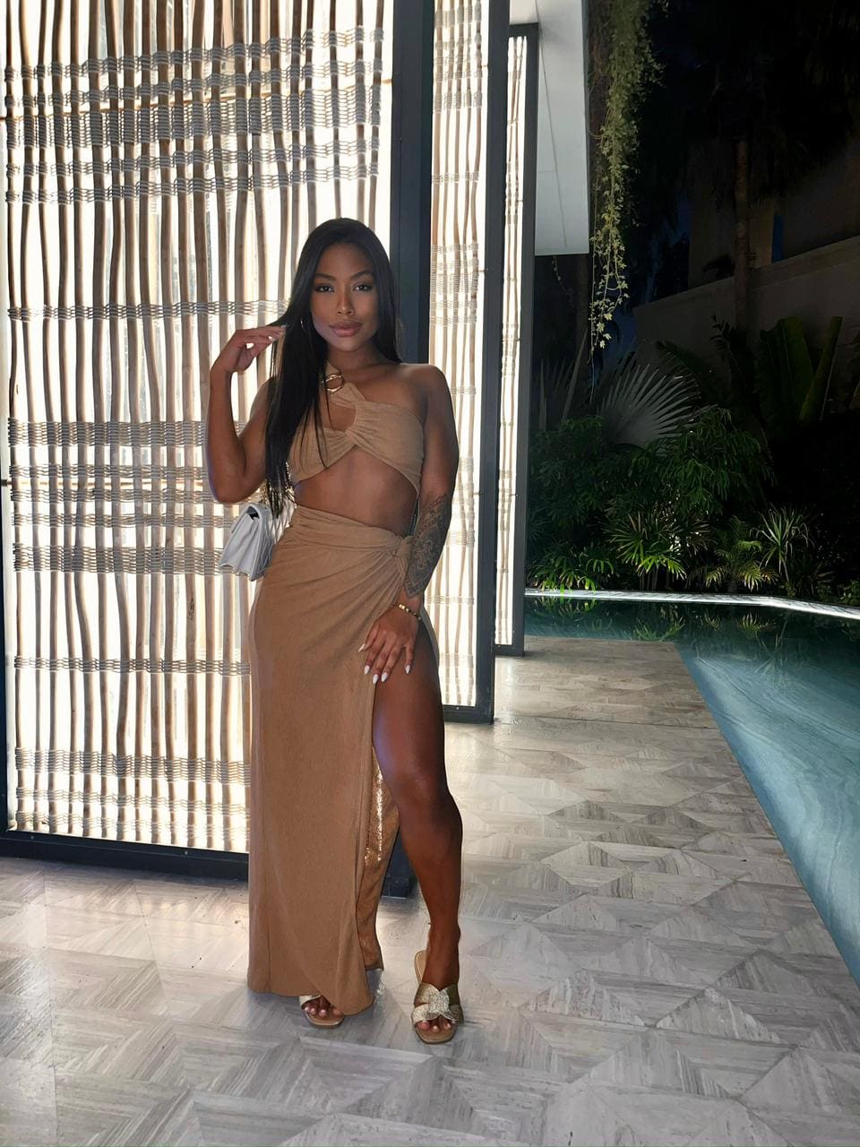 Woman in a tan halter top and high-slit maxi skirt poses by a woven screen and poolside area.