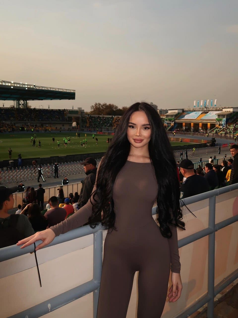 A woman in a brown long-sleeved bodysuit stands at a stadium railing, overlooking a football field and spectators at dusk.