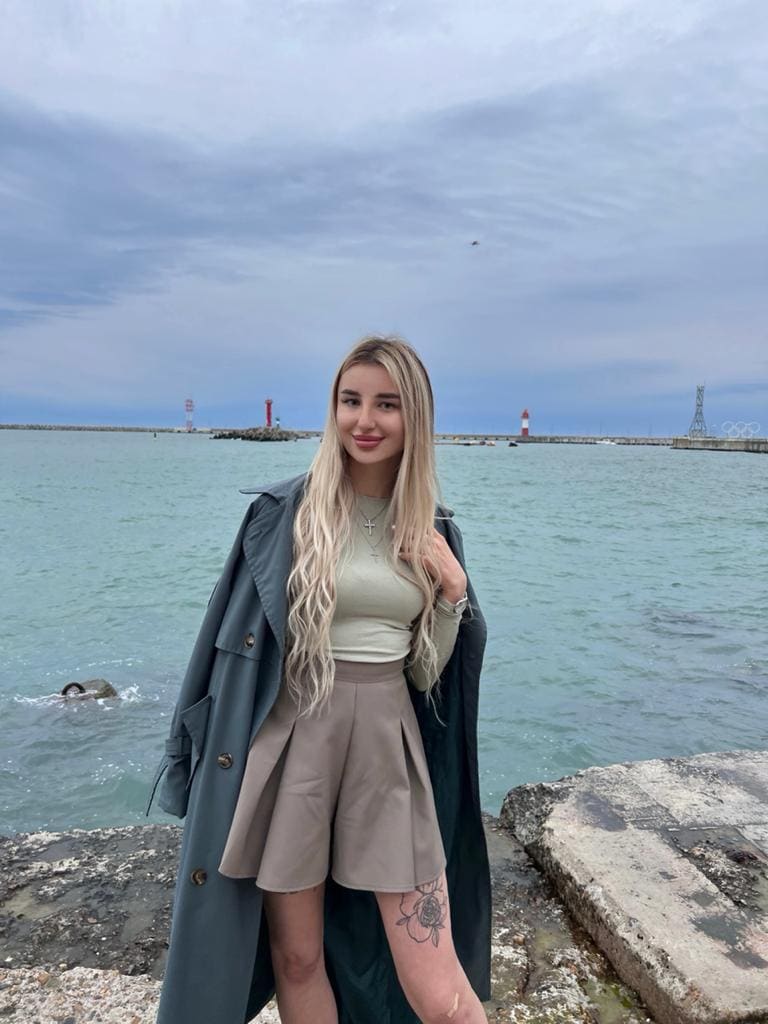 A woman stands by a stone seawall in a light green top, beige skirt, and dark green coat, displaying a rose tattoo on her thigh.