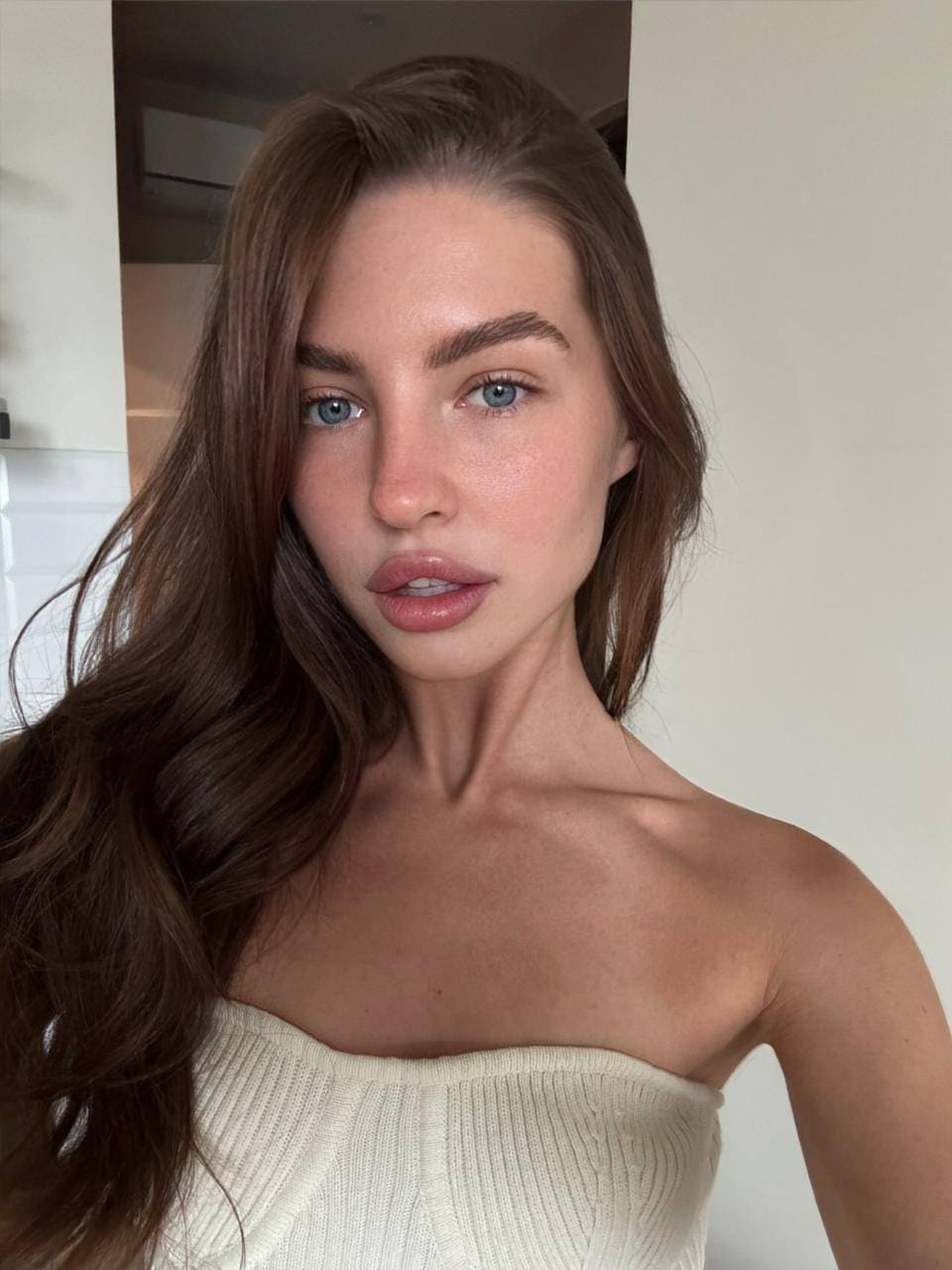 Viollen faces the camera in a cream ribbed strapless top, standing indoors against white walls with a doorway visible behind her.