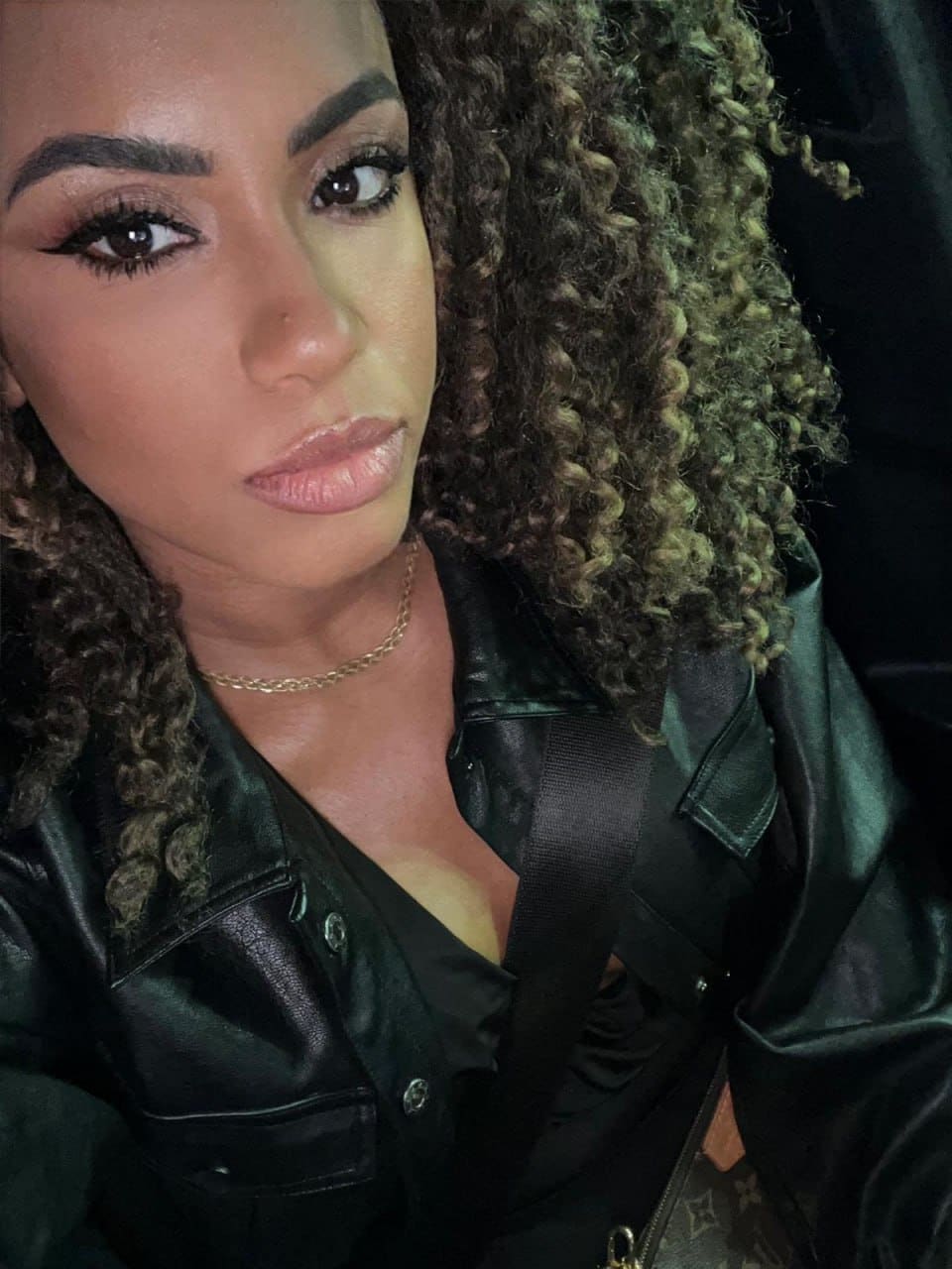 A close-up portrait of a woman in a black leather jacket and gold chain, seated with a seatbelt across her chest against a dark background.