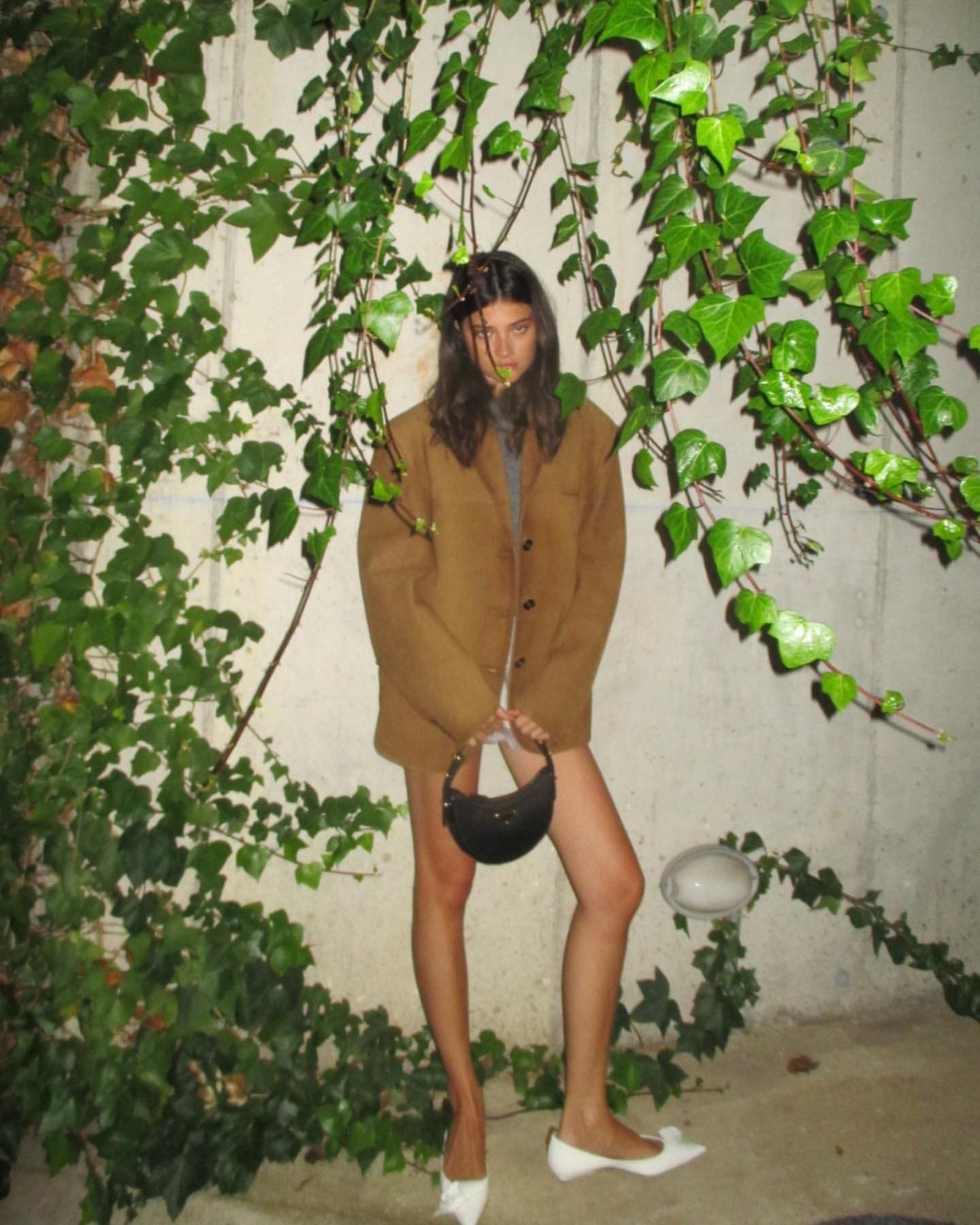 In front of an ivy-covered wall, she wears an oversized brown jacket and holds a small black bag.