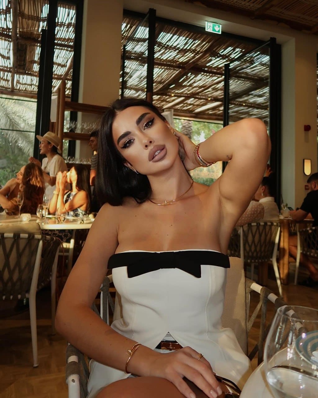 Daniela sits in a restaurant chair wearing a white strapless dress with a black bow, hand raised to her neck, with diners and a thatched roof behind.