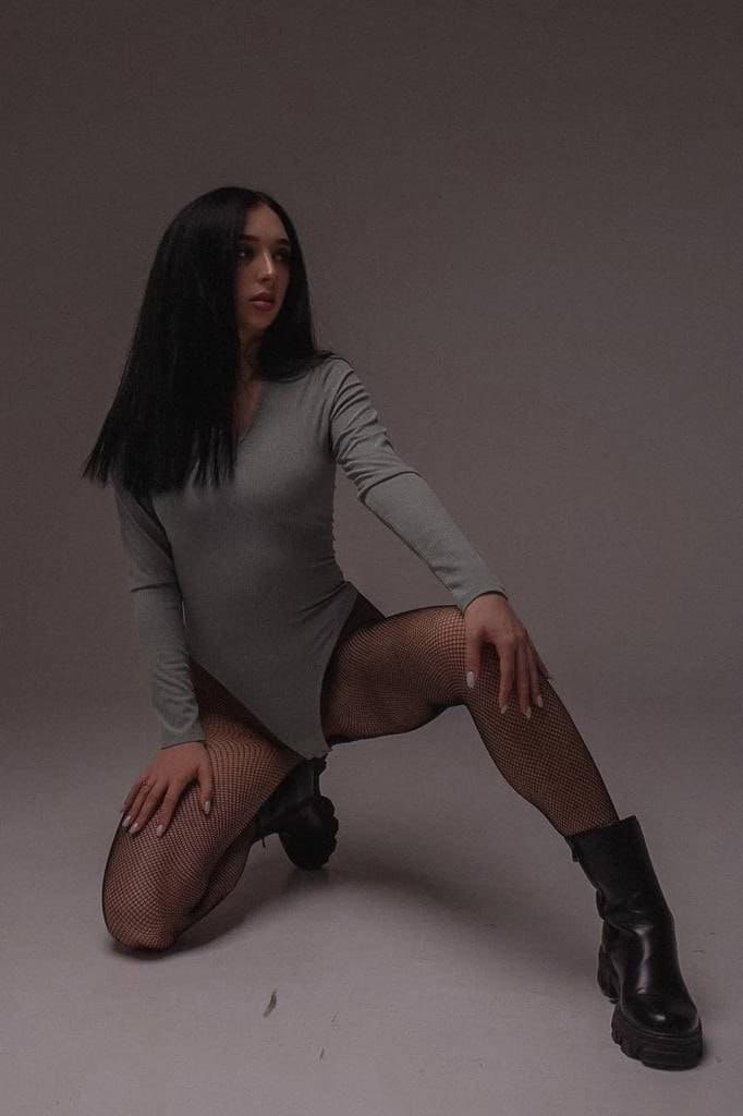 A woman kneels in a studio wearing a grey long-sleeve mini dress, black fishnet stockings, and platform boots, with one hand on her thigh.