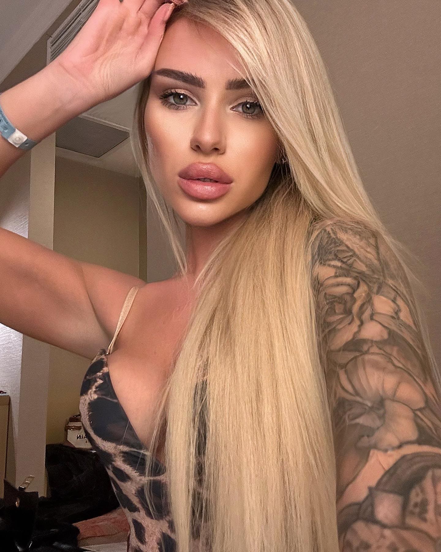 Indoors, she wears a low-cut patterned top with thin straps, raising her right hand to her forehead while showing a floral arm tattoo.