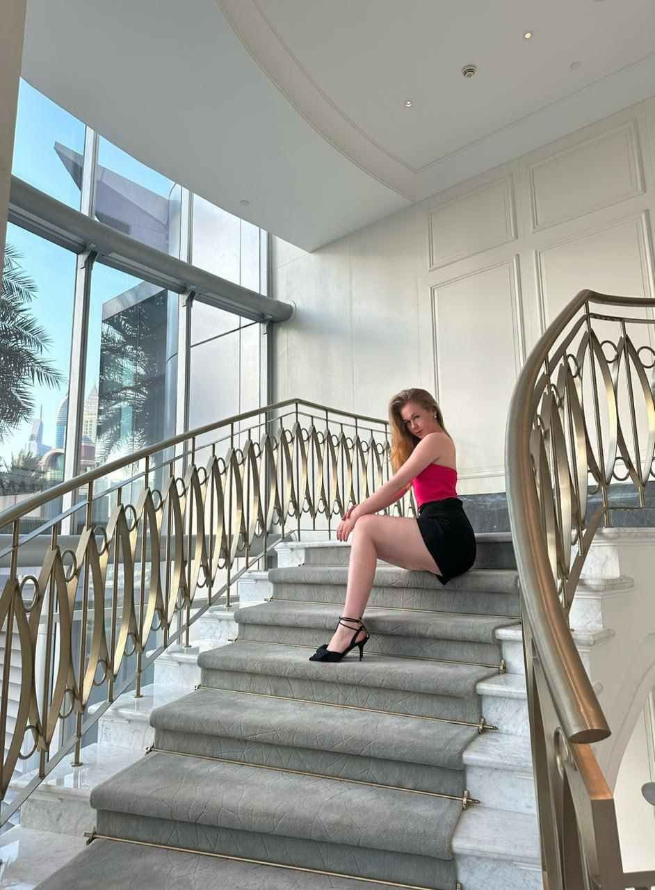 A woman sits on a marble staircase in a pink strapless top, black shorts, and heels, within a bright interior space.