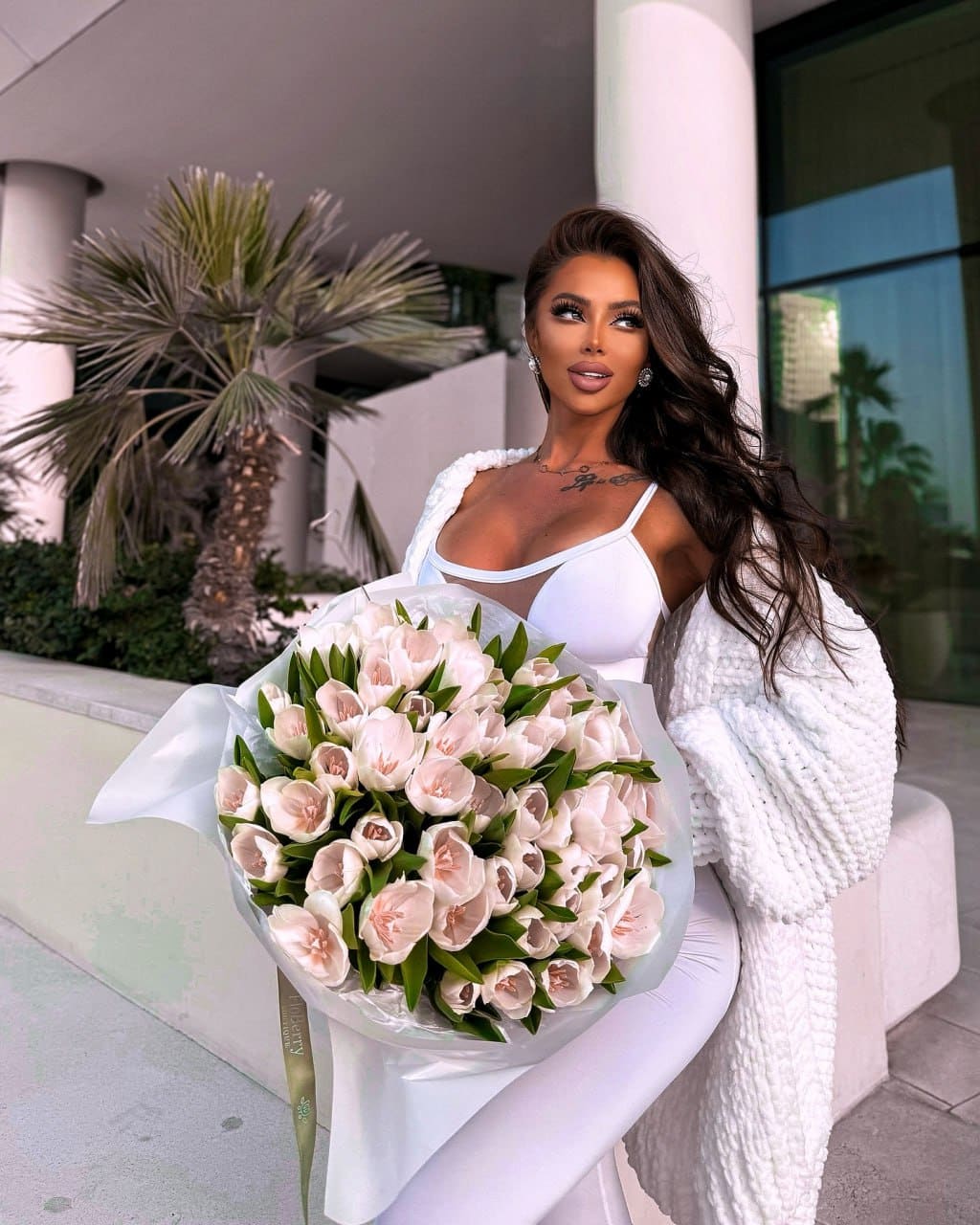 A woman stands outdoors holding pale pink tulips, wearing a white strappy top, leggings, and a textured cardigan, with a palm tree behind her.