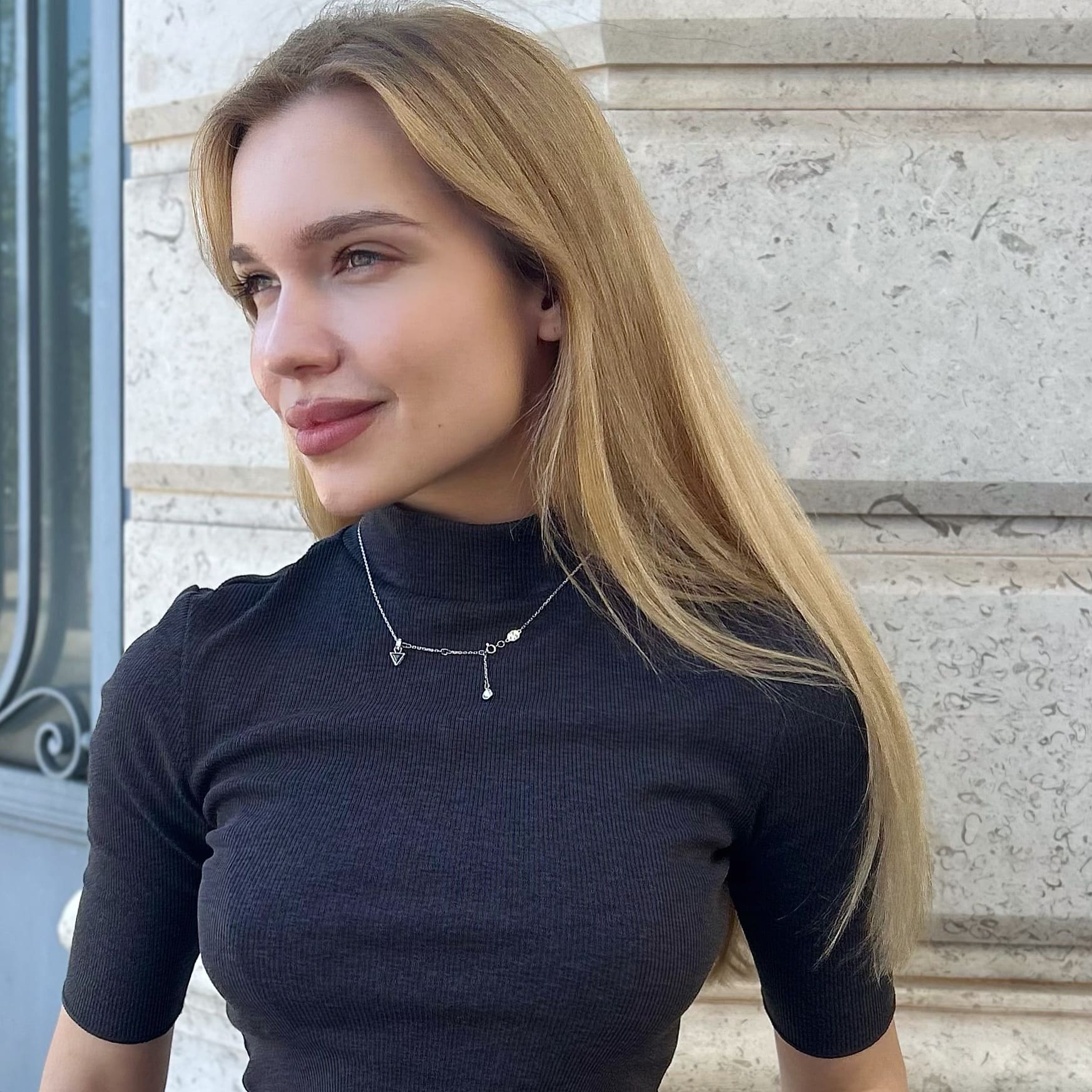 Olya stands before a light stone wall in a fitted black turtleneck and silver charm necklace, head turned slightly with a faint smile.